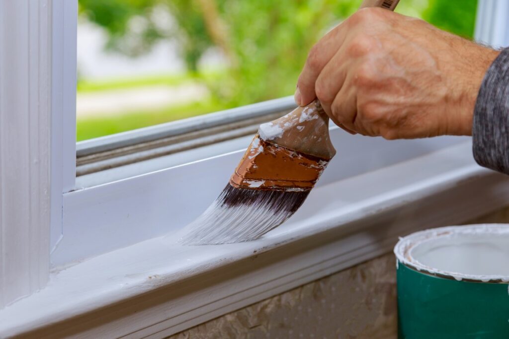 Closeup of a person painting a wooden window trim with white paint.