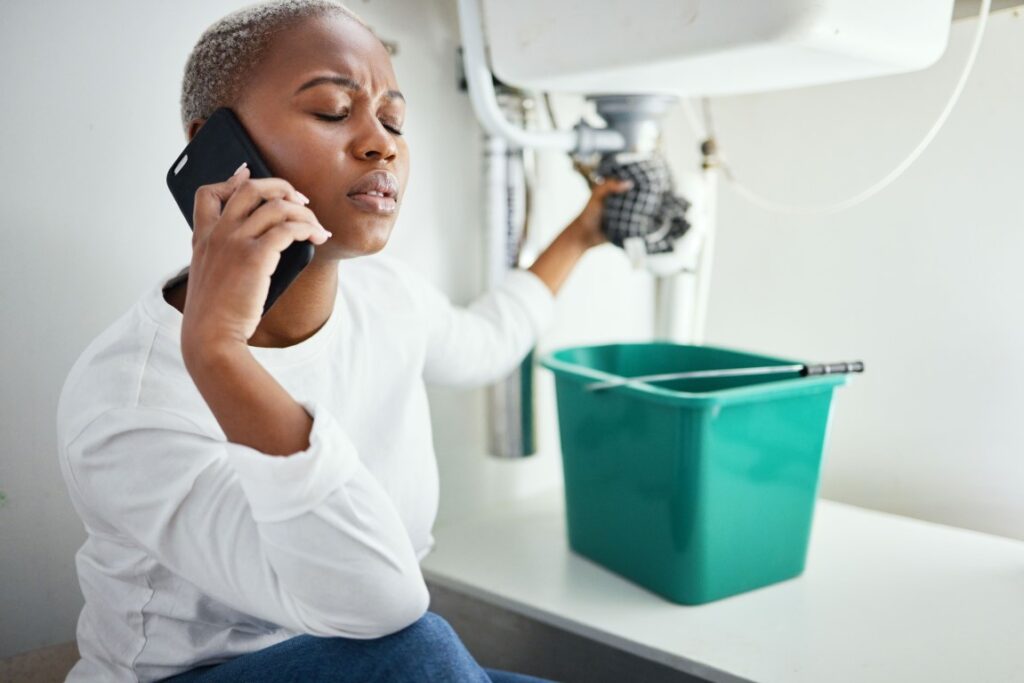 An anxious woman on her cell phone holding a cloth around a pipe under the sink over a bucket.