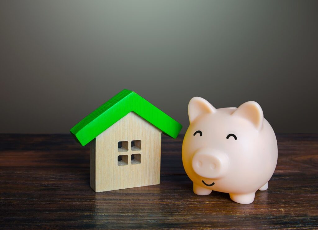 A smiling piggy bank next to a wooden model of a house on a wooden surface.