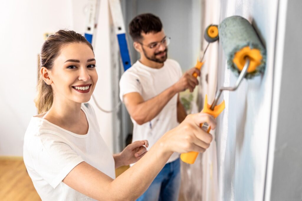 A smiling young couple painting the interior of a home with paint rollers.